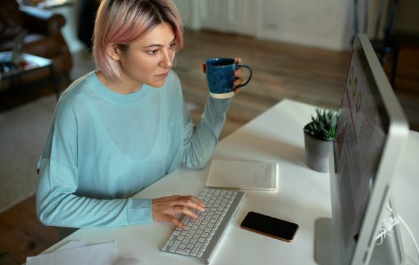 vista-de-alto-angulo-de-uma-jovem-e-atraente-freelancer-com-expressao-facial-concentrada-enquanto-trabalhava-longe-de-casa-sentado-em-frente-ao-computador-desktop-digitando-bebendo-cafe